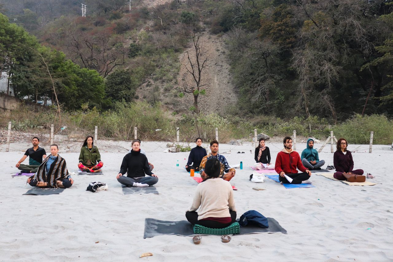 Home A group practicing meditation on a sandy beach in Rishikesh, India, promoting mindfulness and relaxation.